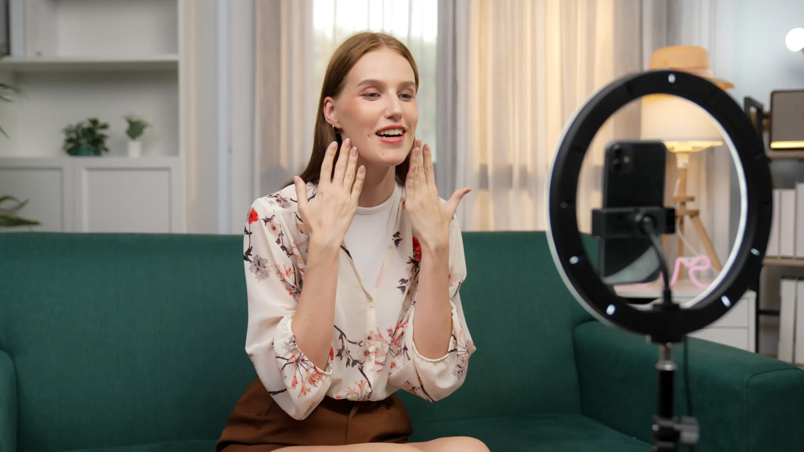 Vlogger woman sits on a green velvet sofa in front of a bright ring light and camera, holding her hands up to her face with a happy, confident expression.